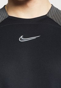 Camisa atlética ligera negra con acentos texturizados en los hombros en gris y un logo blanco de Nike en el pecho. Tejido suave, diseño ajustado.
