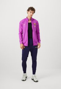 Paarse sportjacket met hoge kraag, geborduurd logo en ritsluiting. Gecombineerd met marineblauwe tapered broek. Witte en grijze sportschoenen maken de look af.