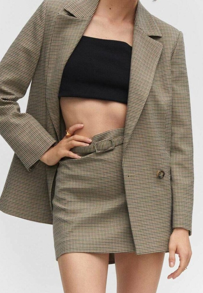 Femme portant un blazer à carreaux beige et une mini-jupe assortie avec ceinture, associée à un top tube noir, main reposant sur la taille.