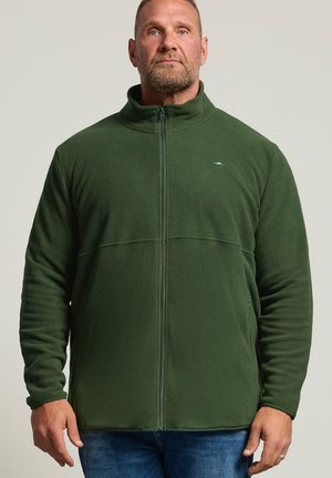 Man met kort haar en baard draagt een groene fleecejas met rits en blauwe spijkerbroek, staand tegen een effen achtergrond.