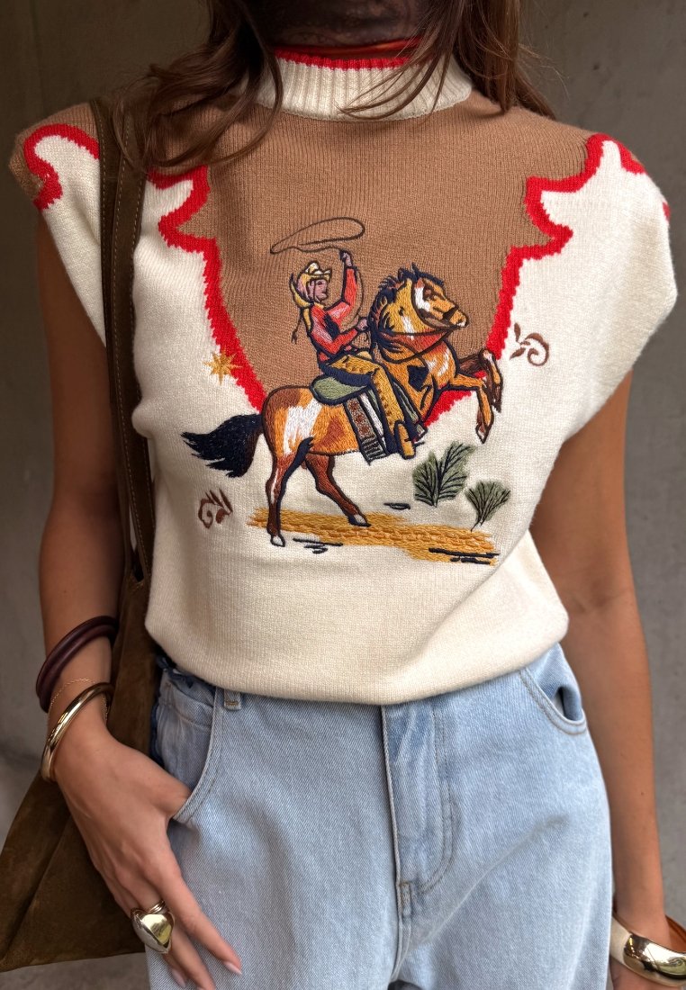 Αμάνικο πλεκτό τοπ με εικονογράφηση καουμπόι, καουμπόι που ιππεύει άλογο που σκαρφαλώνει με λασό· φορεμένο με ανοιχτόχρωμο τζιν και καφέ λουρί τσάντας.