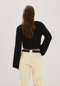 Pull cropped en tricot noir avec manches évasées, associé à un pantalon cargo beige taille haute et une ceinture tressée noire. Détails de conception texturés visibles.