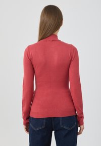 Sweter z długim rękawem, dopasowany, w koralowym kolorze z wysokim dekoltem i prążkowanymi mankietami. Tekstura jest gładka, idealna do nakładania na dżins.