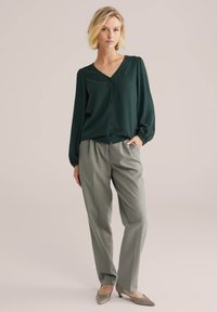 Blusa verde scuro con scollatura a V, maniche lunghe e chiusura a bottoni, abbinata a pantaloni grigi chiari su misura e décolleté beige a punta.