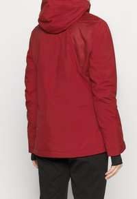Czerwona wodoodporna kurtka z kapturem, charakteryzująca się gładką teksturą i wyrazistym krojem. Zawiera boczne kieszenie oraz kontrastujące panele materiałowe.