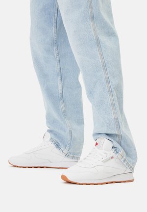 Valged spordijalatsid tekstuurse pealispinnaga, lamedate paelte ja kummist tallaga, kombineeritud hele siniste teksapükstega, millel on nähtav õmblus.