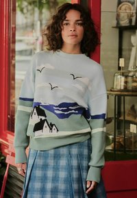 Frau mit lockigem Haar, die einen pastellfarbenen Pullover mit einem Design von einem Küstendorf und Vögeln trägt, kombiniert mit einem blauen Karorock und neben einer roten Tür steht.