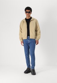 Tan bomber jacket with elastic cuffs, black t-shirt, blue jeans, and black sneakers. Model wearing round sunglasses, standing against a white background.