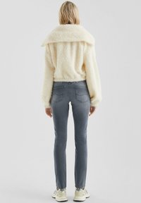Giacca soffice di color crema con ampio colletto, abbinata a jeans grigi aderenti. Il modello indossa scarpe da ginnastica chiare. Nessun motivo o accessorio visibile.