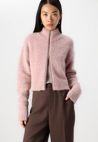 Sudadera corta de color rosa claro con cuello alto y cremallera completa. Fabricada con un material suave y esponjoso, con dobladillo y puños acanalados. Combinada con pantalones marrones.