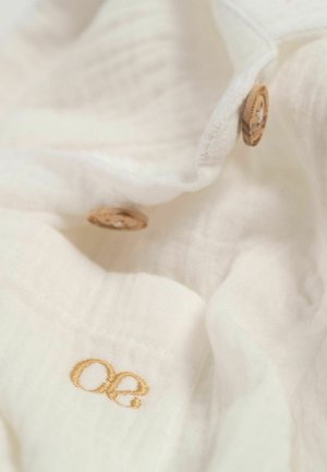 Tissu blanc avec une apparence douce et texturée. Présente des boutons en bois et une broderie dorée affichant les initiales "oe."