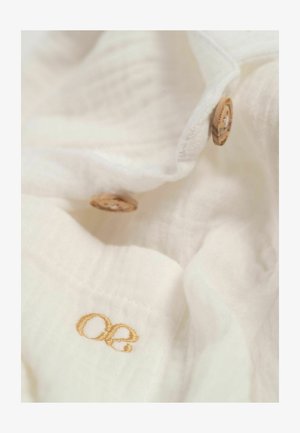 Tissu blanc avec une apparence douce et texturée. Présente des boutons en bois et une broderie dorée affichant les initiales "oe."