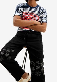 Pasiasty granatowo-biały t-shirt z czerwonym wzorem graficznym; czarne spodnie z haftem kwiatowym na dolnej części nogawek; jasne buty.