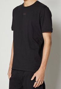 T-shirt en coton noir avec des manches courtes et un col rond. Patch logo discret sur la poitrine. Texture lisse, coupe décontractée.