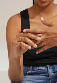 Gold rings in various designs, including twisted and geometric shapes, worn on hands with natural nails against a dark top.