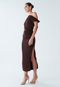 Donna che indossa un vestito midi marrone con spalle scoperte, arricciato, con spacco sulla coscia e sandali dorati con tacco alto e cinturini, in piedi davanti a uno sfondo semplice.