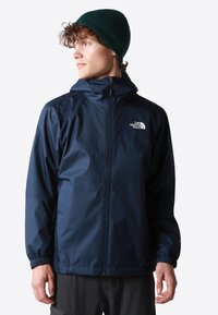 Chaqueta impermeable azul marino con capucha, puños elásticos y cremallera frontal. Presenta el logo blanco en el lado izquierdo del pecho.