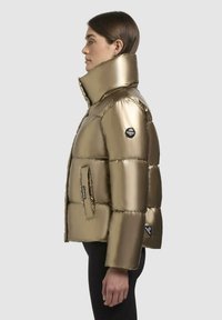 Metalická zlatá péřová bunda s vysokým límcem, zipem vpředu a oversized prošívaným designem. Obsahuje nášivku s logem na rukávu a boční kapsy.