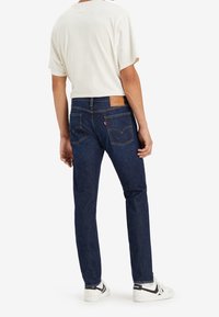 Mörkblå denimjeans med rak passform, läder bakpatch och två bakfickor. Bärs med en vit kortärmad skjorta och sneakers.