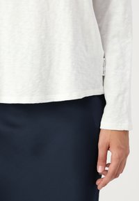 White long-sleeve shirt with a relaxed fit and subtle textured fabric, paired with a navy blue bottom, showing a hand at the edge.