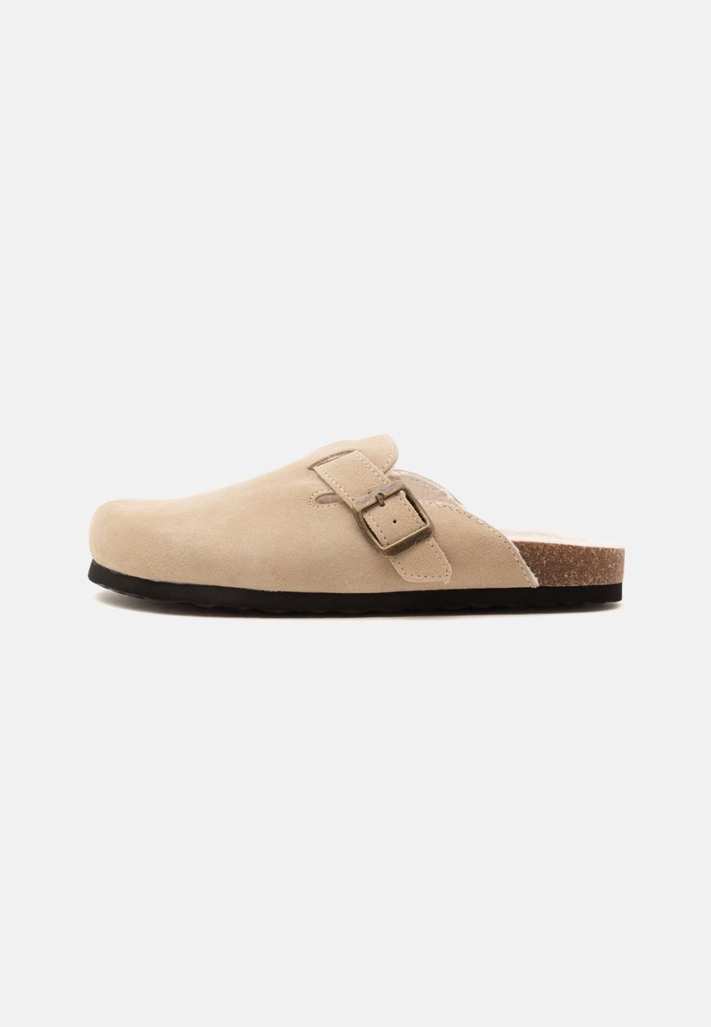 Μπεζ παπούτσι τύπου slip-on με κλειστή μύτη, ρυθμιζόμενο λουράκι με αγκράφα, πάτο από φελλό και μαύρη σόλα, εμφανιζόμενο σε πλάγια όψη σε λευκό φόντο.