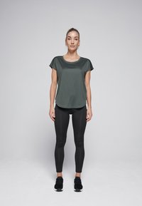 Donkergroene shirt met korte mouwen en een ronde hals, gemaakt van textuurstof, gecombineerd met zwarte leggings en zwarte sportschoenen.