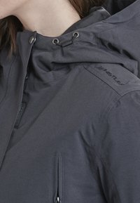 Veste à capuche grise avec une surface texturée, dotée de quincaillerie en métal, de cordons de serrage réglables et d'une poche latérale zippée.