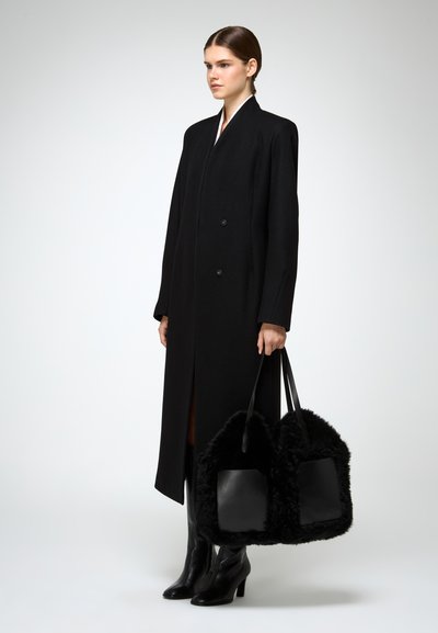 Cappotto lungo nero in lana con doppi bottoni e spacco laterale. La modella tiene una borsa nera in pelliccia sintetica con dettagli in pelle. Stivali neri lucidi con tacco.