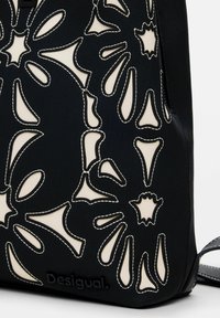 Schwarze Stofftasche mit ausgestanztem floralen Design, das einen cremefarbenen Hintergrund sichtbar macht. Sie verfügt über weiße Nähte und der Markenname "Desigual" ist eingeprägt.