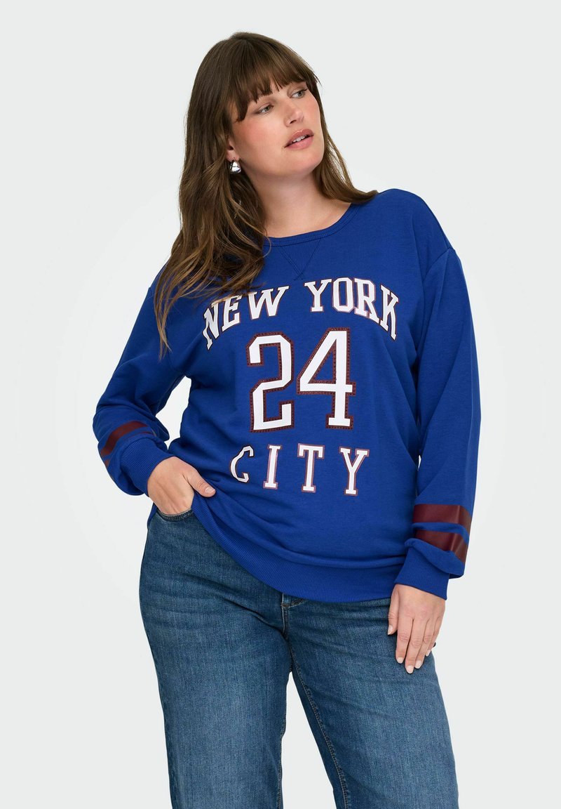 Preveliki plavi dukserica s otisom "NEW YORK 24 CITY" u bijeloj i burgundiji. Ima dugačke rukave s burgundi prugama. Materijal je od mješavine pamuka.