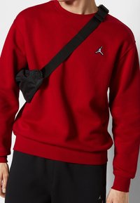 Sudadera roja con cuello redondo, puños acanalados y un pequeño logo negro de Jordan. Se lleva un bolso negro al hombro sujeto con una correa.