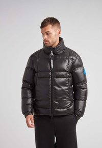 Sort pufferjakke med blank overflade, høj krave, lynlåslukning og blå logoakcent. Tyk quiltet design med elastiske manchetter.