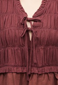 Κοντινό πλάνο ενδύματος από υφή υφάσματος μπορντό με ραφές που δημιουργούν πτυχές και δύο δεμένα φιόγκους στον εμπρόσθιο λαιμό.