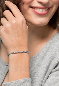 Silver chain bracelet with rectangular links, worn on a wrist. The wearer has light skin and is smiling, dressed in a gray sweater.