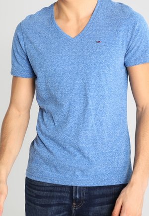 Uomo che indossa una maglietta blu aderente a maniche corte con scollo a V e jeans blu scuro, in piedi davanti a uno sfondo chiaro e uniforme.