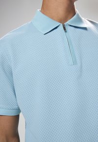 Polo shirt azzurro chiaro con un motivo a quadri testurizzato, maniche corte e chiusura a mezzo zip con zip a contrasto.