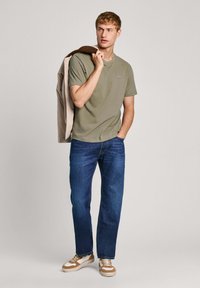 T-shirt in cotone verde oliva, vestibilità rilassata, abbinata a jeans di denim blu. La modella tiene una giacca leggera e indossa sneakers beige e marroni.