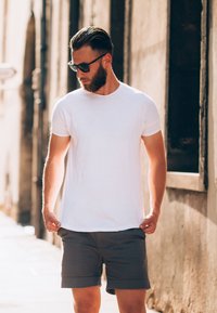 T-shirt bianca, maniche corte, collo rotondo. Pantaloni corti grigi, lunghezza sopra il ginocchio. Occhiali da sole, ambientazione urbana informale con muri di pietra.