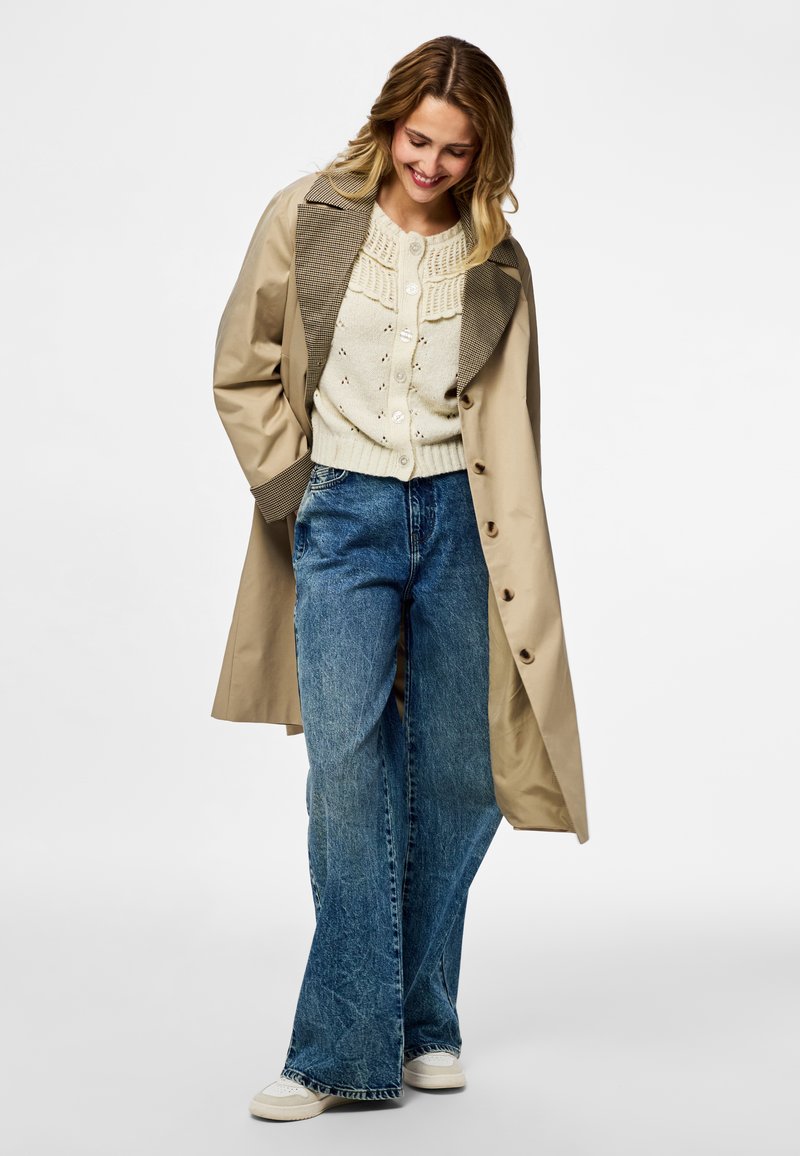 Femme portant un trench beige sur un cardigan en maille crème, jean large bleu et baskets blanches, regardant vers le bas et souriant.
