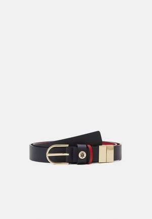 Ceinture en cuir noir avec boucle dorée, passant rouge et emblème rond doré avec des initiales sur un fond clair uni.