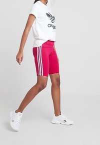 Rosa sportshorts med tre vita ränder på varje sida, parat med en vit t-shirt med en svart logotyp, och vita sneakers.