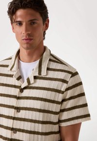 Camisa de manga corta con un diseño de punto texturizado, que presenta una tela crema con rayas verde olivo y botones marrones en la tapeta delantera.