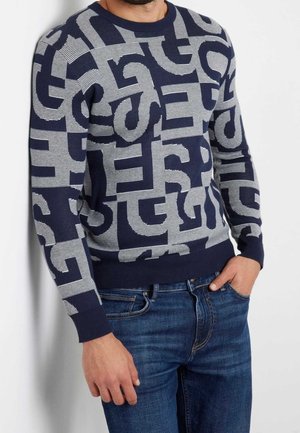 Uomo con maglione blu scuro con audaci motivi astratti di lettere in grigio chiaro, abbinato a jeans blu, in piedi contro un muro bianco.