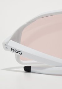 Lunettes de soleil blanches avec une monture légère et des verres teintés en rose. Elles présentent un logo sur le côté et une extrémité noire texturée sur la branche.