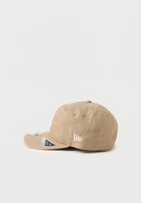 Šviesiai rudos spalvos kepurė su išlenktu šoniniu kraštu, tekstūruota medžiaga, balta siuvinėta logo ir reguliuojama nugaros juosta. Etiketės pritvirtintos po kraštu.