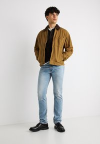 Νέος άνδρας που στέκεται με τα χέρια στις τσέπες, φοράει καφέ μπουφάν, μαύρο πουκάμισο, ανοιχτό μπλε τζιν και μαύρα παπούτσια, πάνω σε απλό λευκό φόντο.