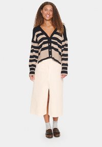 Cardigan court rayé noir et beige avec des boutons, associé à une jupe midi crème avec une fente avant et des chaussures marron.