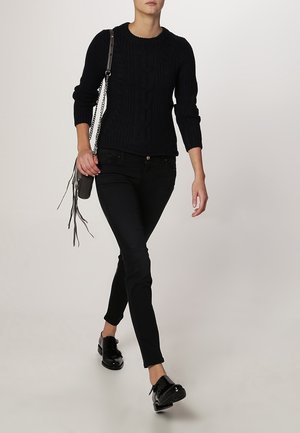 Mujer caminando con suéter negro de punto trenzado, pantalones pitillo negros, zapatos negros brillantes con cordones y llevando un bolso de hombro gris con flecos.