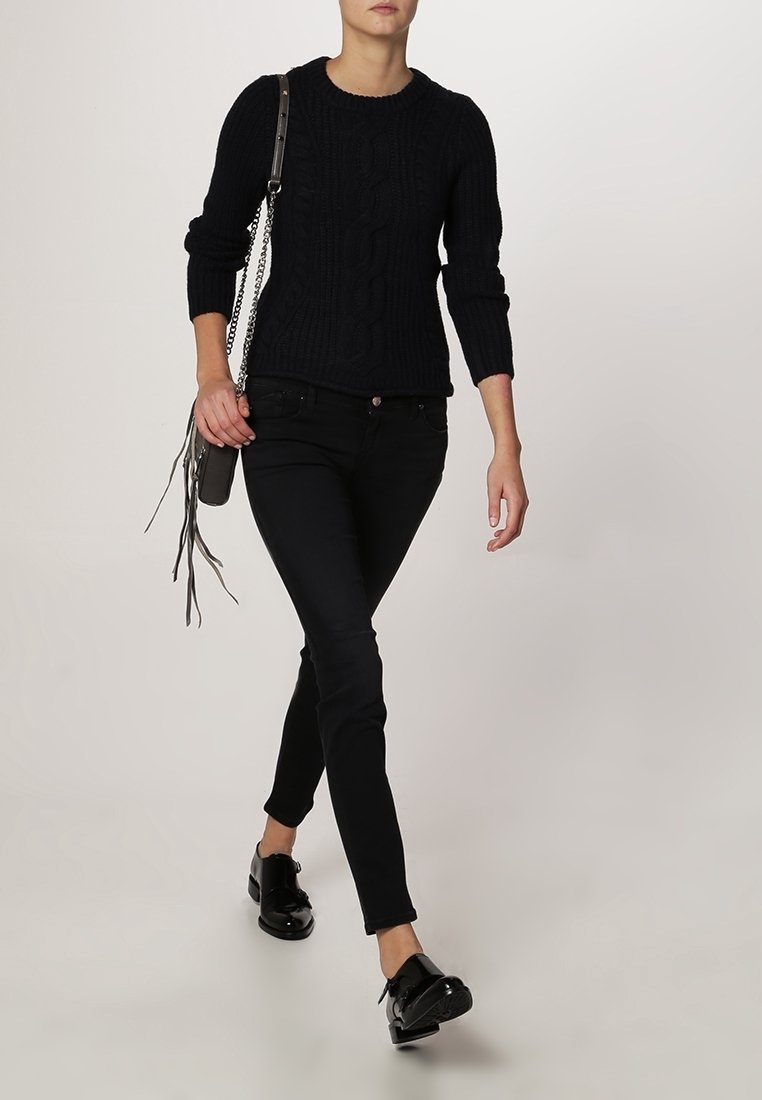 Femme marchant vêtue d'un pull noir à torsades, d'un pantalon skinny noir, de chaussures noires brillantes à lacets, et portant un sac à bandoulière gris à franges.