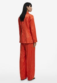 Traje de satén rojo con bordados florales; americana ajustada con mangas largas y pantalones de pierna ancha. Textura suave y diseño estructurado.